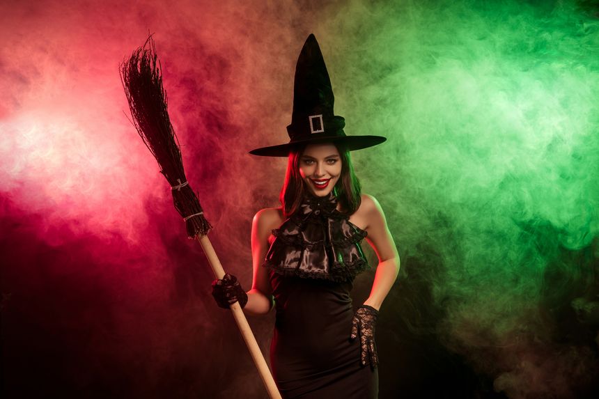 Captivating Halloween imagery featuring a beautiful witch in elegant attire holding a broomstick, surrounded by colorful, mystical fog.